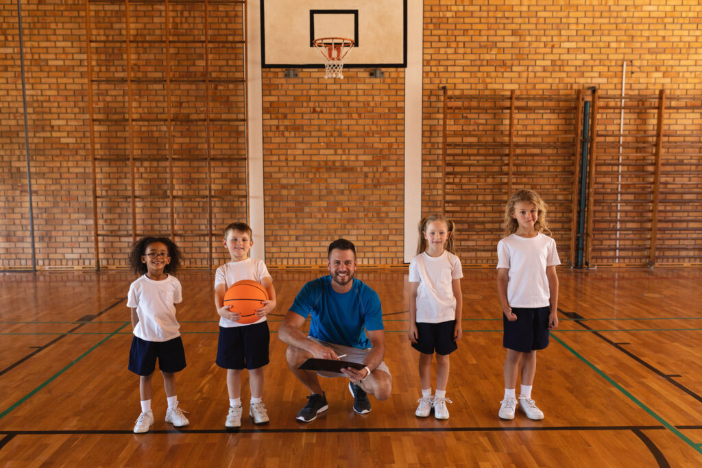 Front view of happy basketball coach and schoolkids looking at camera at basketball court